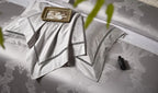 Folded silver gray bedding with layered damask shams, tonal couture trim, and a platinum satin sheen – L&B