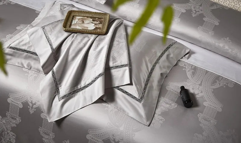 Folded silver gray bedding with layered damask shams, tonal couture trim, and a platinum satin sheen – L&B